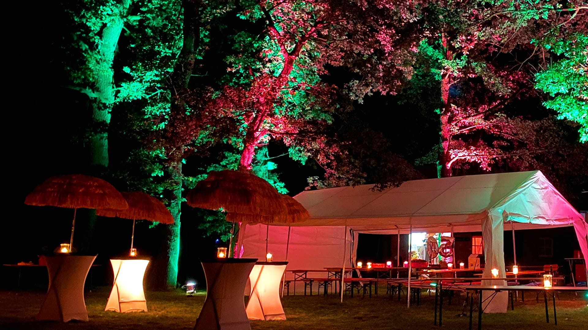 Festlich beleuchtetes Partyzelt bei Nacht im Garten, umgeben von Bäumen mit grüner und roter Lichtinszenierung, davor Stehtische mit Strohschirmen und Kerzenlicht