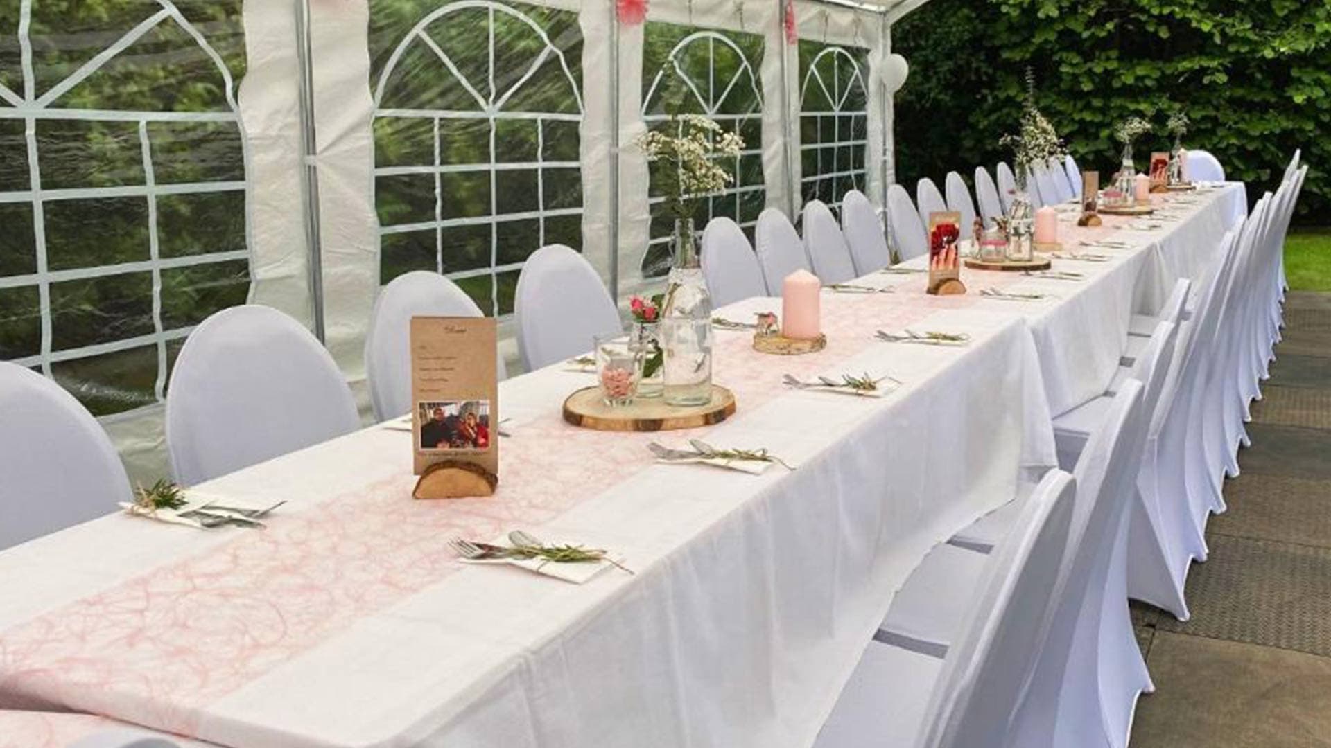 Festlich gedeckte Tafel in einem Pavillonzelt mit weißen Stuhlhussen, Tischdeko und Blumen für eine Hochzeitsfeier